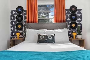 Ein Bett mit grauer Kopfteil, weißen Kissen, einer blauen Decke und einem Kissen mit einem Vinyl-Schallplatten-Design. Zwei Nachttische mit Lampen und einer Topfpflanze sind vorhanden.