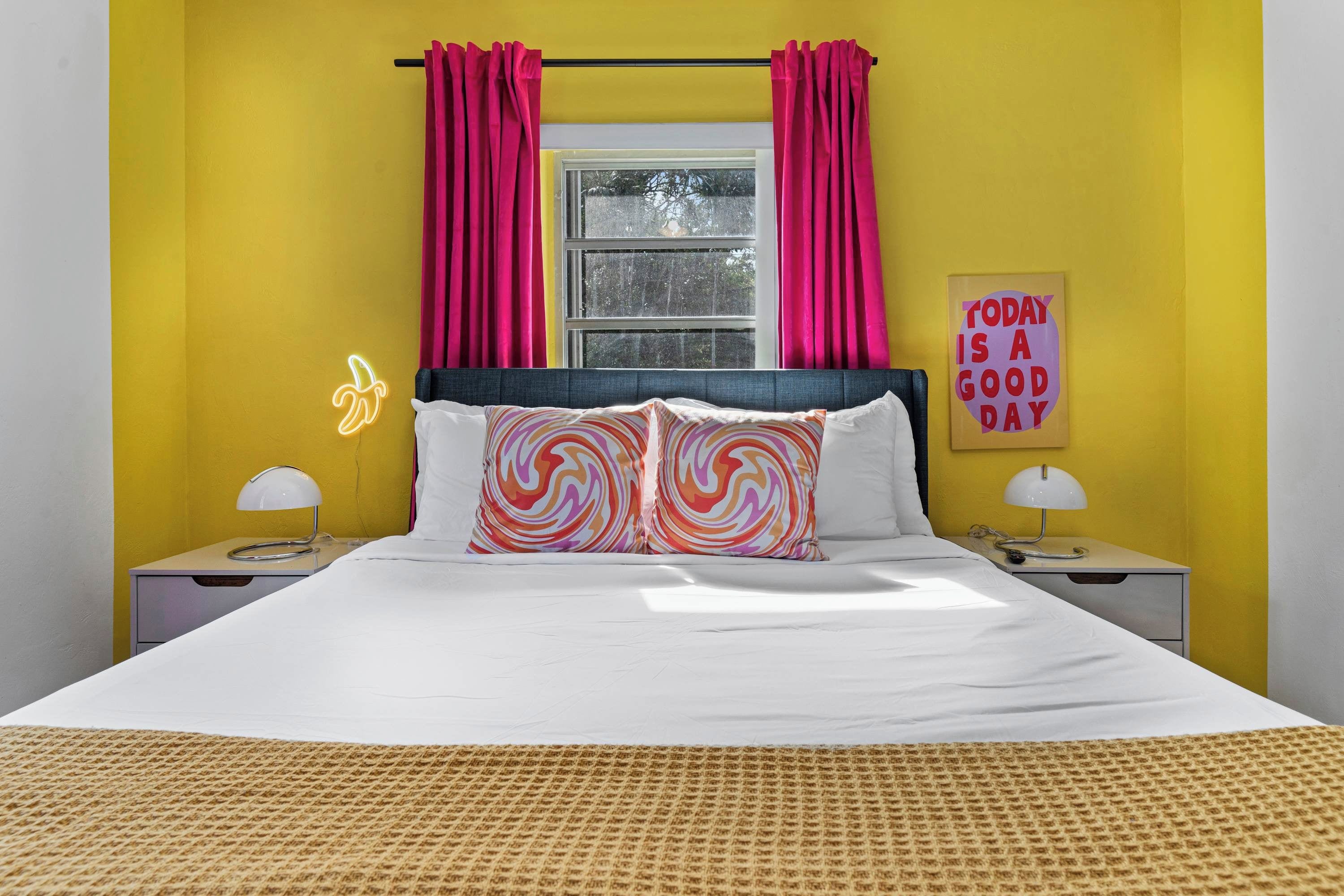 Uma cama com roupa de cama branca, duas mesas de cabeceira, uma janela com cortinas cor-de-rosa e um letreiro com a frase 'Today is a good day'.