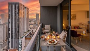 Varanda com mesa de jantar posta para dois, com vista para um horizonte urbano ao pôr do sol.