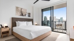 Quarto moderno com uma cama grande, uma mesa de cabeceira com um telefone, uma obra de arte na parede e vista da cidade através de uma porta deslizante.