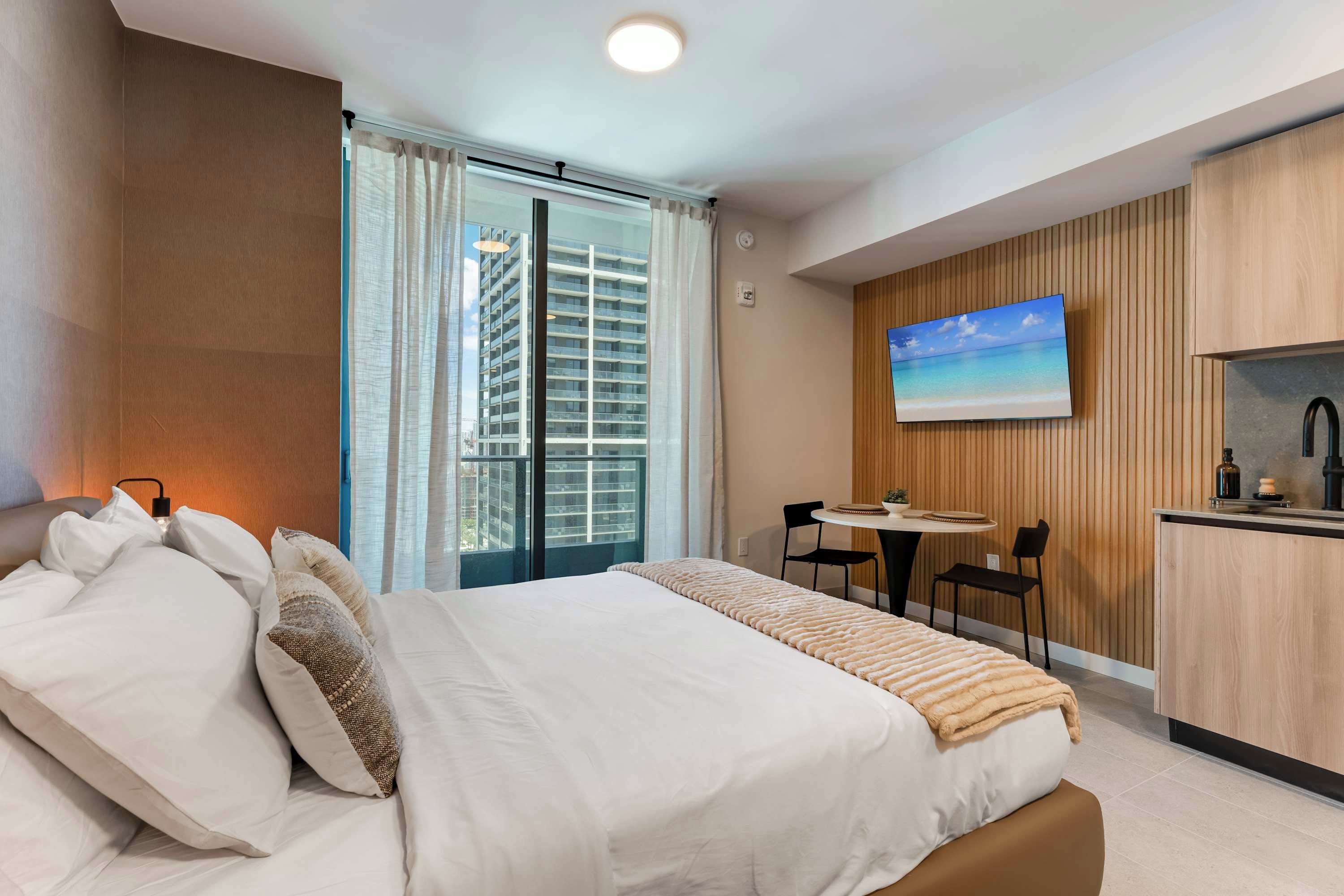 Ein Hotelzimmer mit Bett, Fernseher, einer kleinen Küchenzeile und einem Fenster mit Blick auf ein Hochhaus.