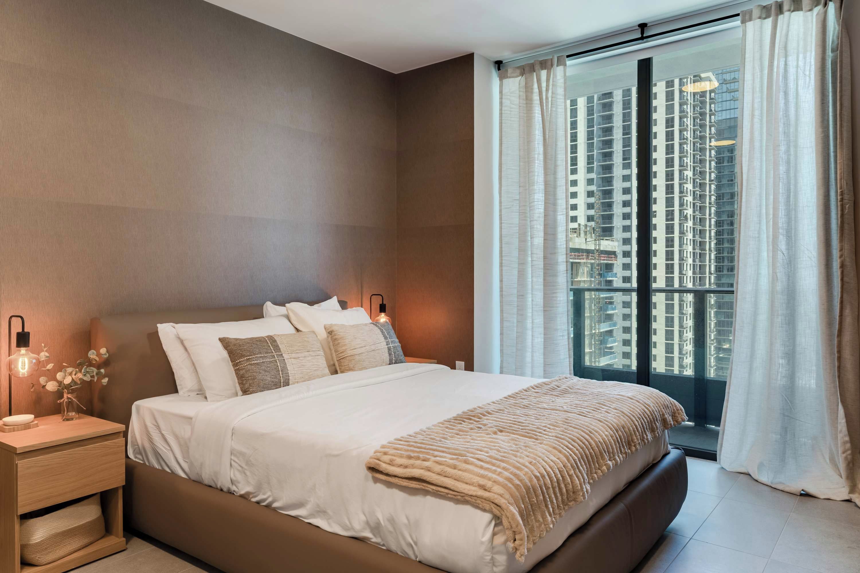Ein modernes Schlafzimmer mit einem großen Bett, Nachttischlampen, einem Beistelltisch und Blick auf hohe Gebäude durch das Fenster.