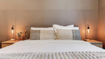 En pænt redt seng med et vamset tæppe, to sengeborde med lamper, og en lampe på væggen.