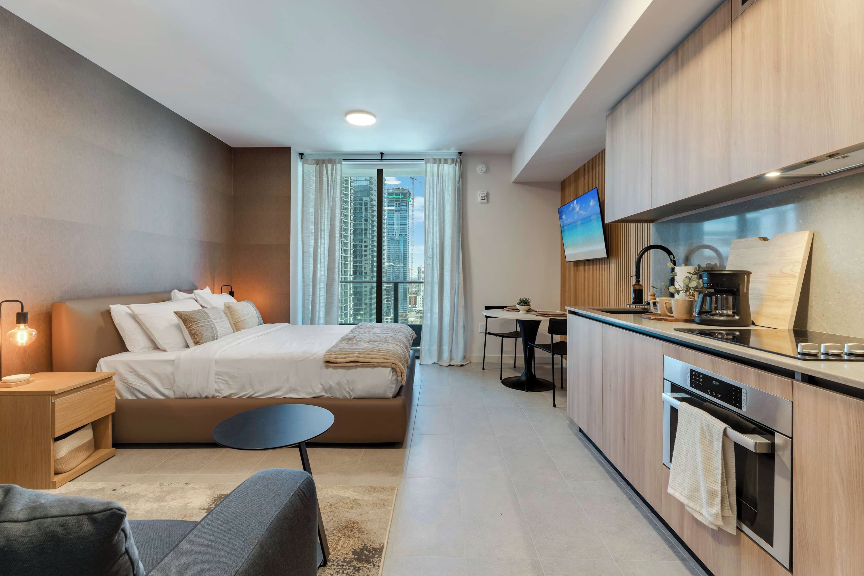 Una habitación de hotel moderna con cama, mesita de noche, silla, kitchenette y televisor.