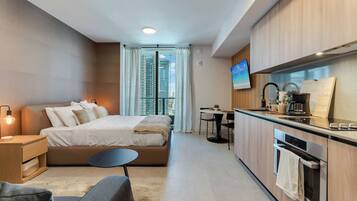 Una habitación de hotel moderna con cama, mesita de noche, silla, kitchenette y televisor.