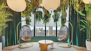 A room with a round wooden table, two wicker chairs, hanging plants, and a view of the city through large windows. - Aura Studio, Heart of Miami With Resort Amenities (Miami)
