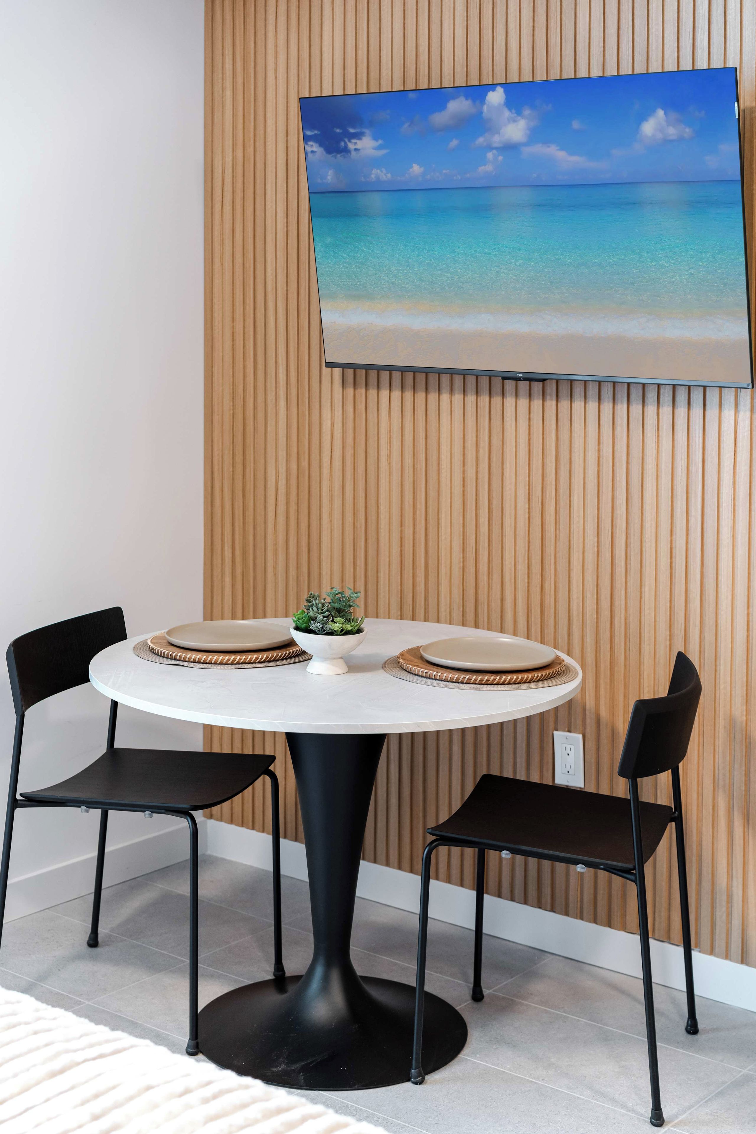 Ein runder Tisch mit zwei Stühlen, einer kleinen Pflanze und einem an der Wand montierten Fernseher, der eine Strandansicht zeigt.