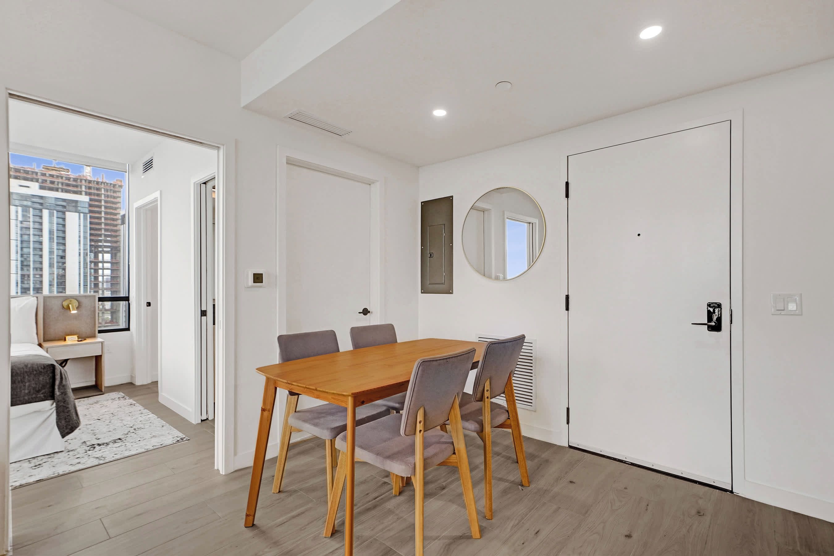 Une chambre moderne avec une table à manger en bois, des chaises grises, un miroir rond et un lit donnant sur la ville.