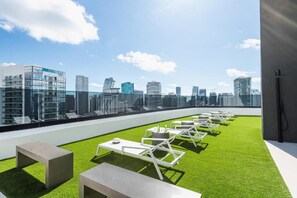 A rooftop terrace with artificial grass, white lounge chairs, and tables, offering a city skyline view. - City Dreams Sauna, Rooftop Pool & Parking (Miami)