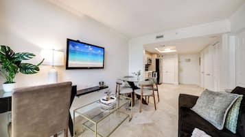 A modern living room with a glass coffee table, a brown leather sofa, a TV mounted on the wall, and a dining area with a round table and chairs.