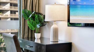 A hotel room with a desk, a potted plant, a lamp, and a TV displaying a beach scene.