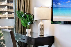 Ein Hotelzimmer mit Schreibtisch, einem Blumentopf, einer Lampe und einem Fernseher, der eine Strandansicht zeigt.