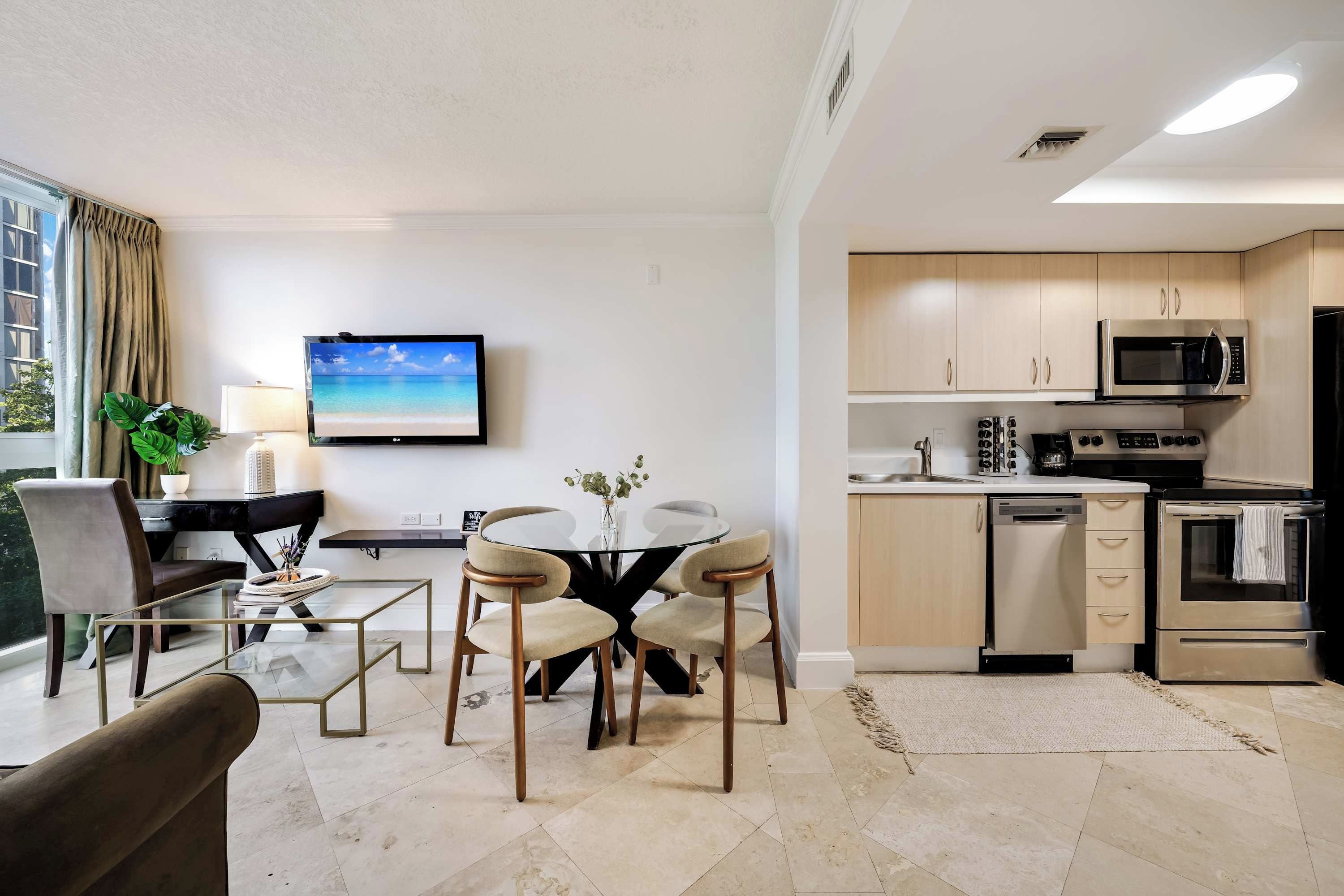 A modern living room with a glass coffee table, two chairs, a TV, and a kitchen with a microwave and oven.