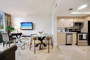 Un salon moderne avec une table basse en verre, deux fauteuils, un téléviseur et une cuisine équipée d’un four à micro-ondes et d’un four traditionnel.