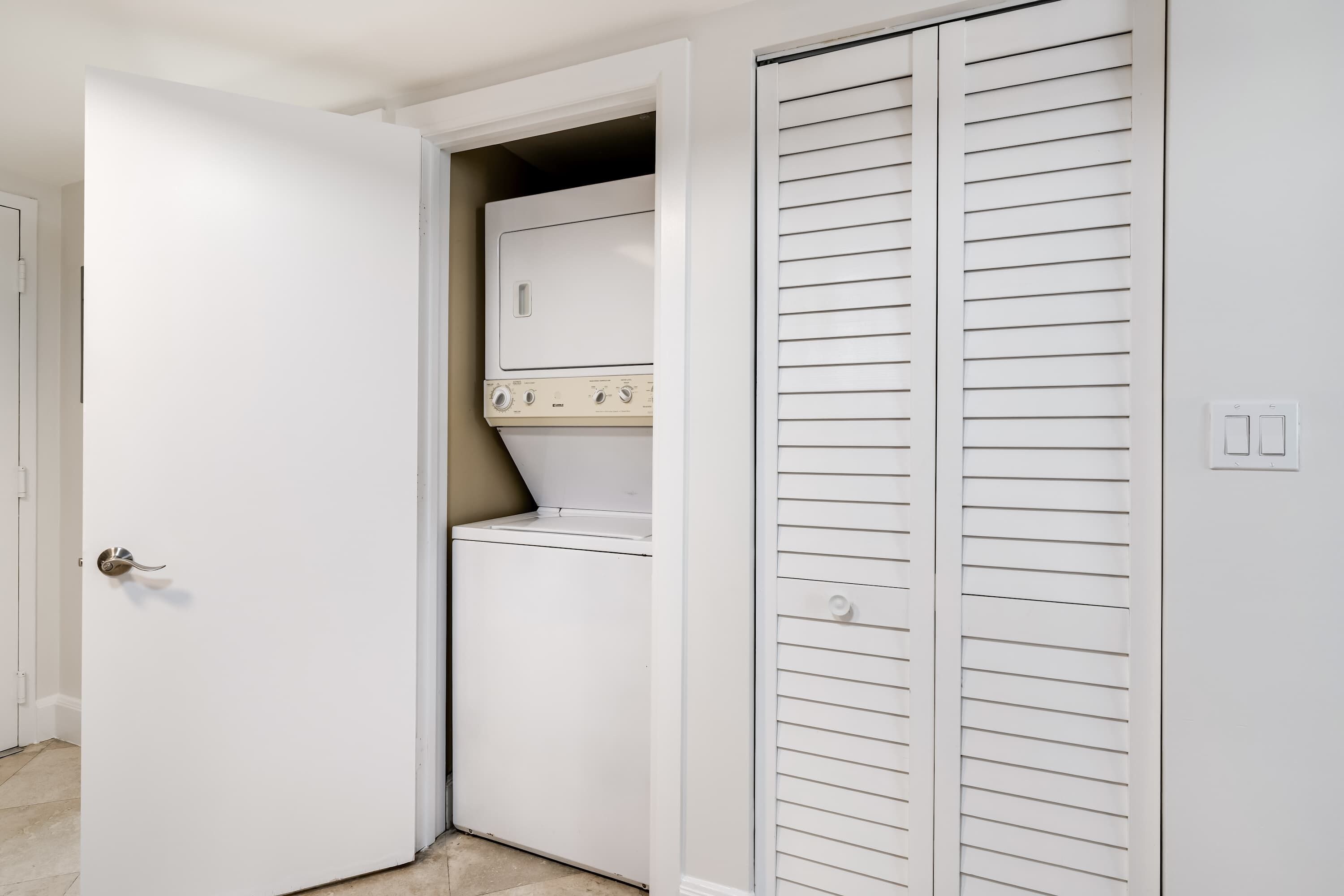 Un cuarto de lavandería con lavadora y secadora, una puerta blanca y un armario blanco con puertas de listones horizontales.