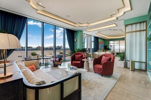 Um lobby moderno de hotel com um sofá, poltronas, uma mesa de centro e grandes janelas com vista para a cidade.