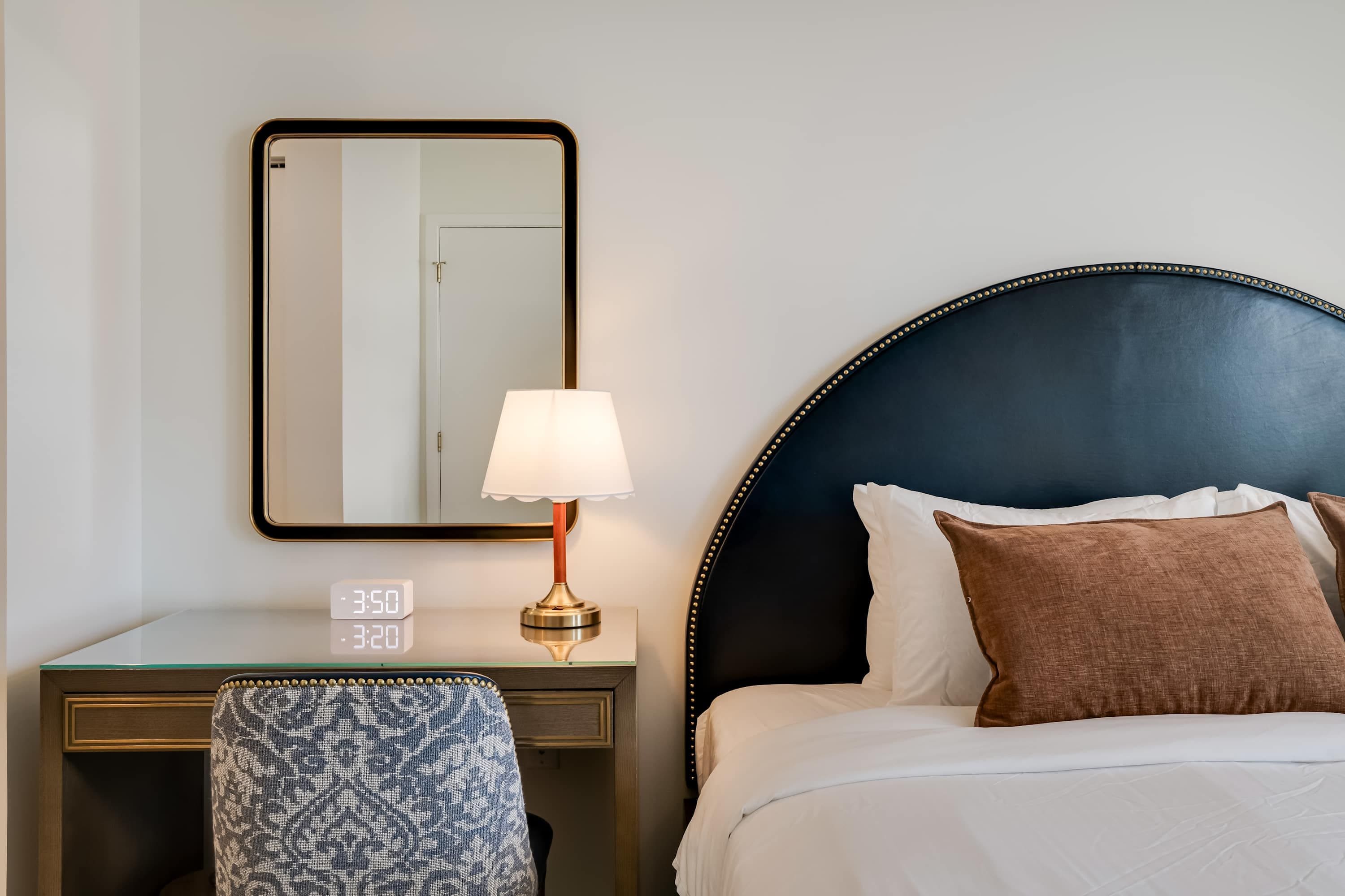 Habitación de hotel con cama, mesita de noche con lámpara, una silla, un espejo y un reloj digital.
