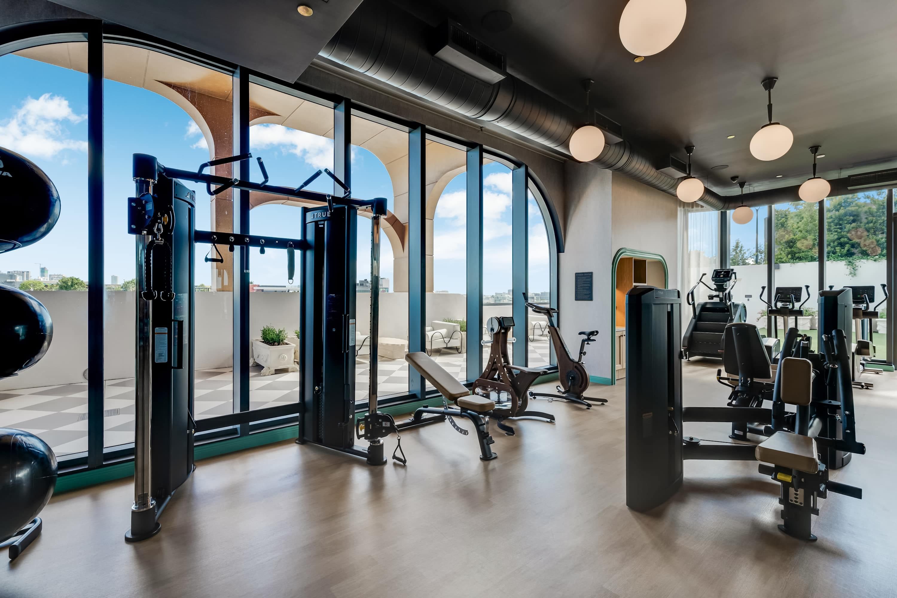 Un gimnasio con equipo de ejercicio, ventanas amplias y vista a la ciudad.