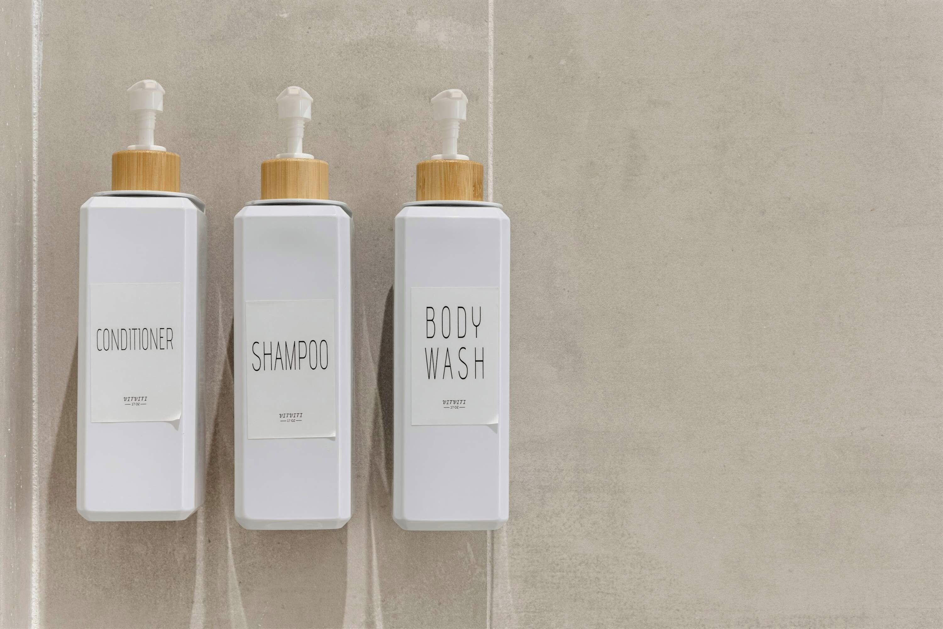 Drie witte flessen met houten dopjes, gelabeld met conditioner, shampoo en douchegel.