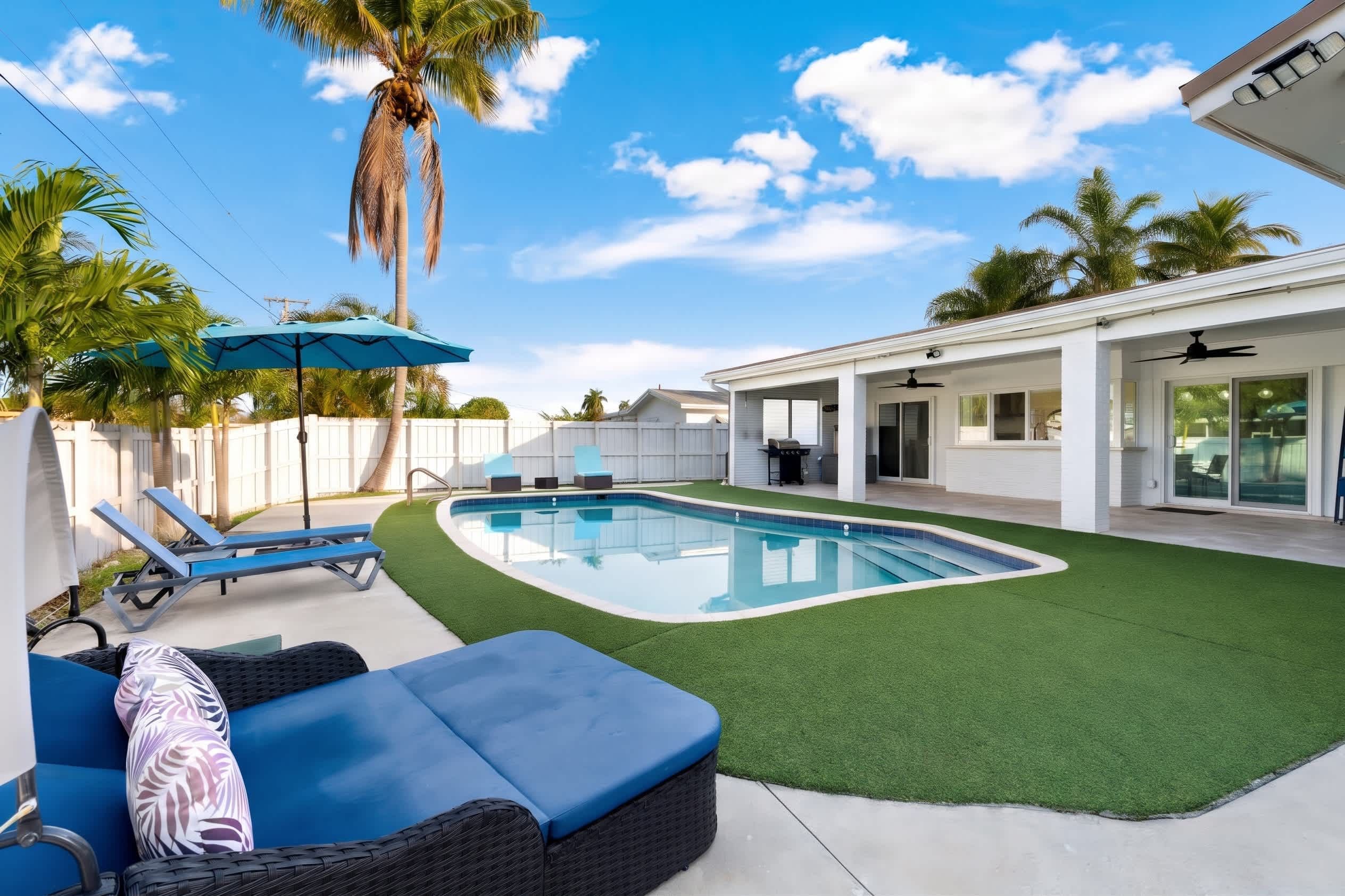 A backyard with a swimming pool, artificial grass, lounge chairs, and a covered patio area.