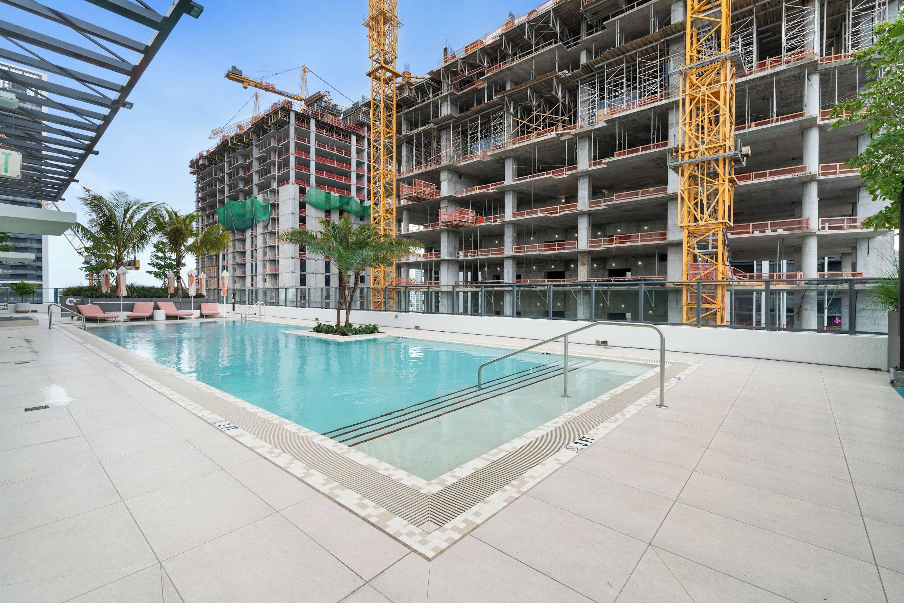 Une piscine dotée d’une terrasse carrelée, entourée d’un bâtiment en construction, composé de plusieurs étages et entouré d’échafaudages.