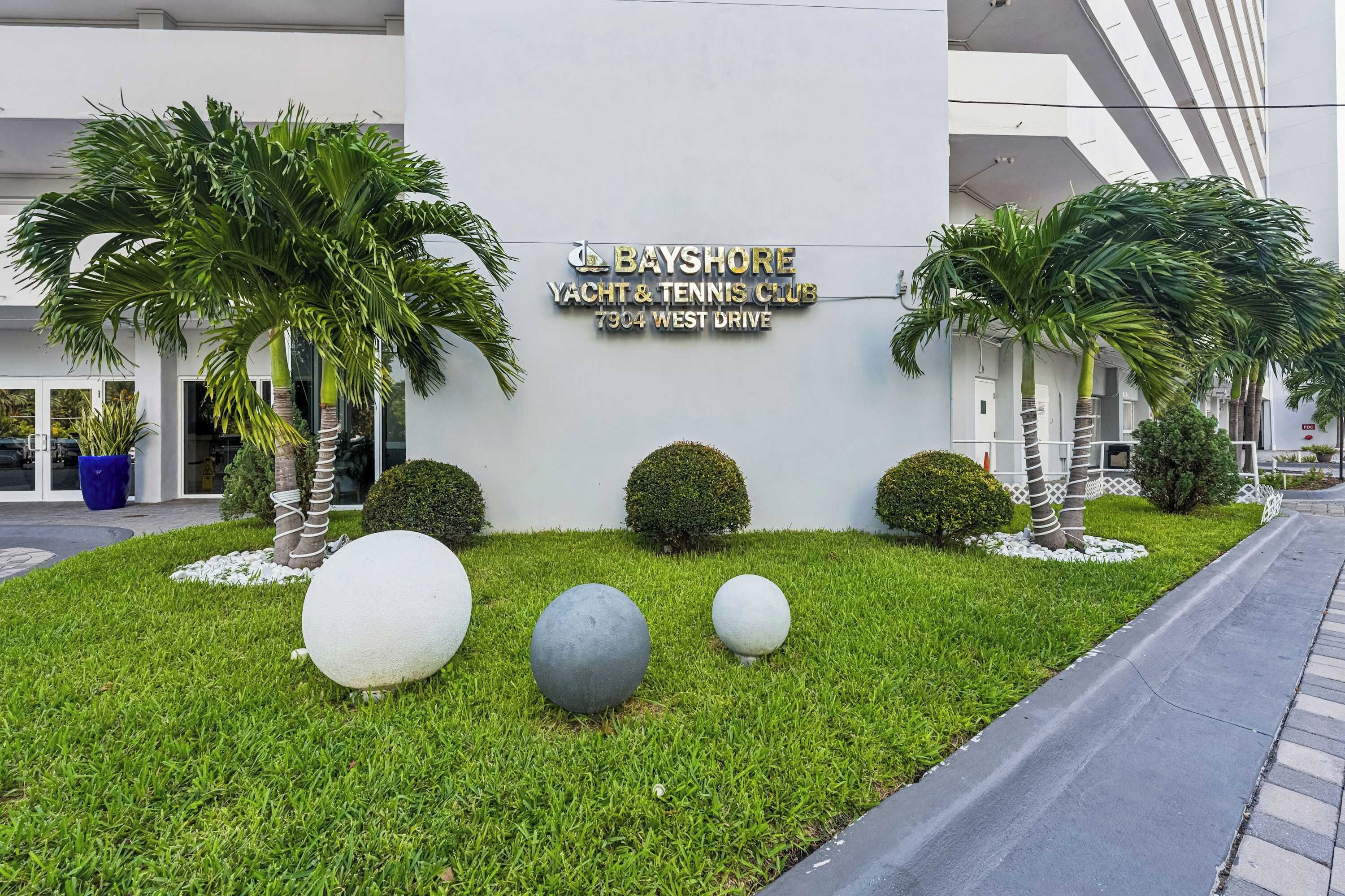 Een bordje voor de Bayshore Yacht & Tennis Club aan 7904 West Drive, omgeven door goed onderhouden gras, palmbomen en bolvormige sculpturen.