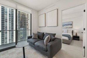 Ein modernes Schlafzimmer mit grauem Sofa, einem kleinen runden Tisch, einem Bett mit Nachttisch und einem großen Fenster mit Blick auf die Stadt.