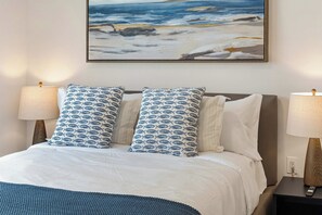 Ein Bett mit weißer Bettdecke und blau gemusterten Kissen, darüber ein Gemälde mit Strandmotiv, und zwei Nachttischlampen auf Holznachtkästen.