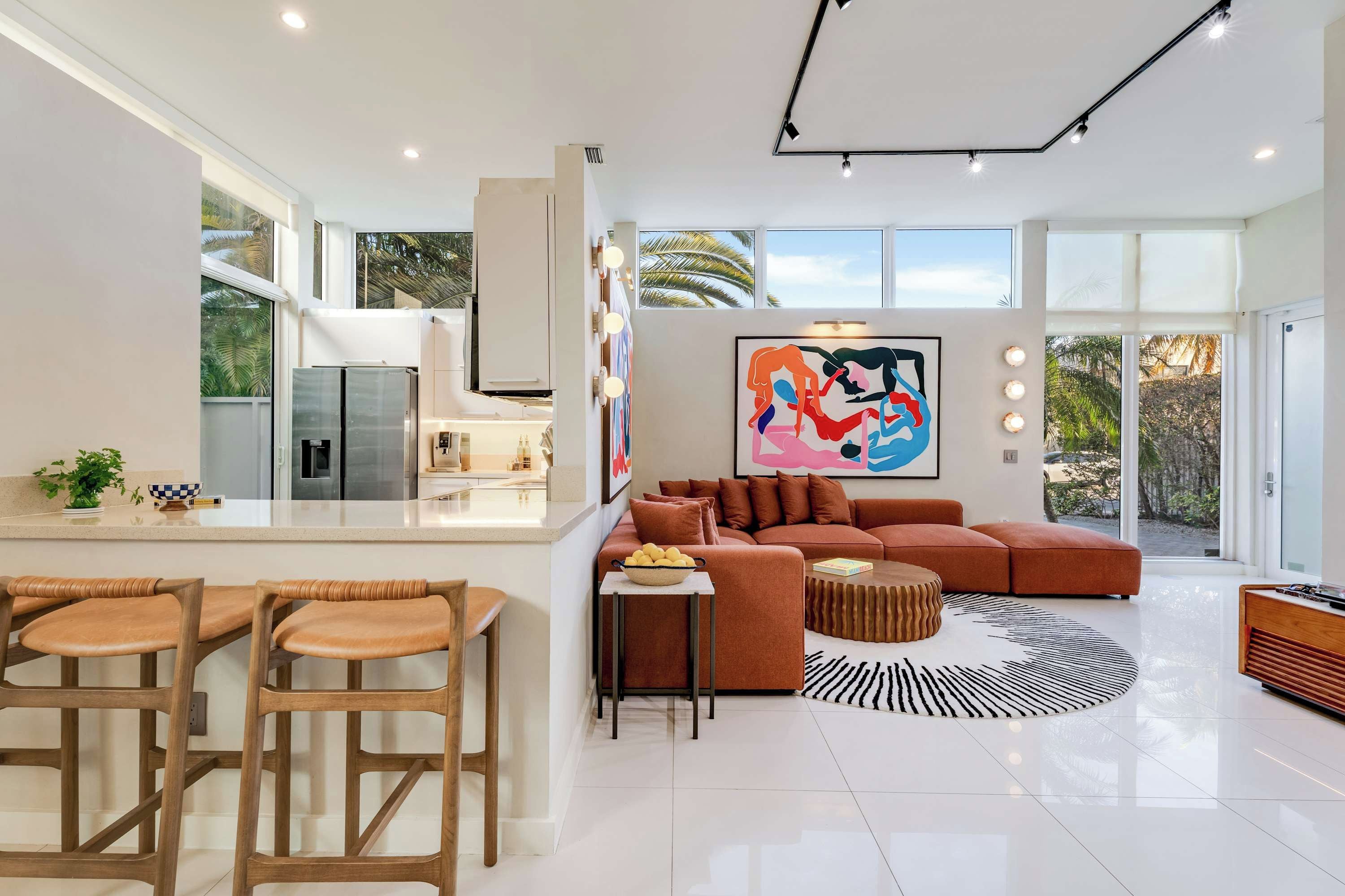 Una cocina moderna con una isla central, un área de comedor con sillas de madera, un sofá grande y un cuadro abstracto colorido.