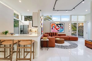 Una cocina moderna con una isla central, un área de comedor con sillas de madera, un sofá grande y un cuadro abstracto colorido.