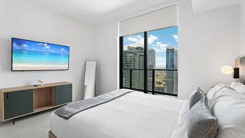 Un dormitorio moderno con cama, televisor, mesita de noche y un amplio ventanal con vistas a la ciudad.