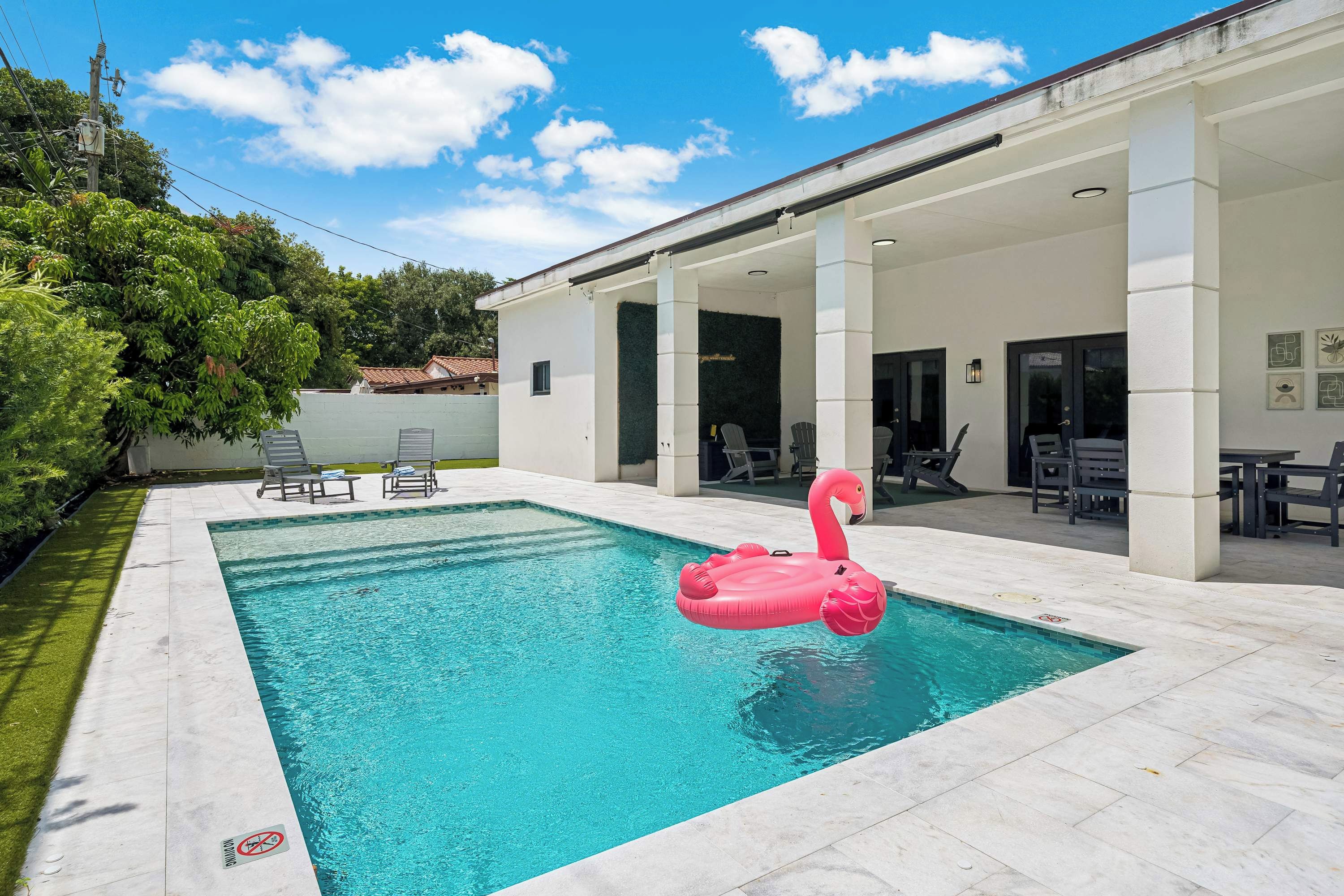 A swimming pool with a pink flamingo float, surrounded by a covered patio area with chairs and tables.