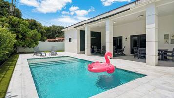 A swimming pool with a pink flamingo float, surrounded by a covered patio area with chairs and tables.