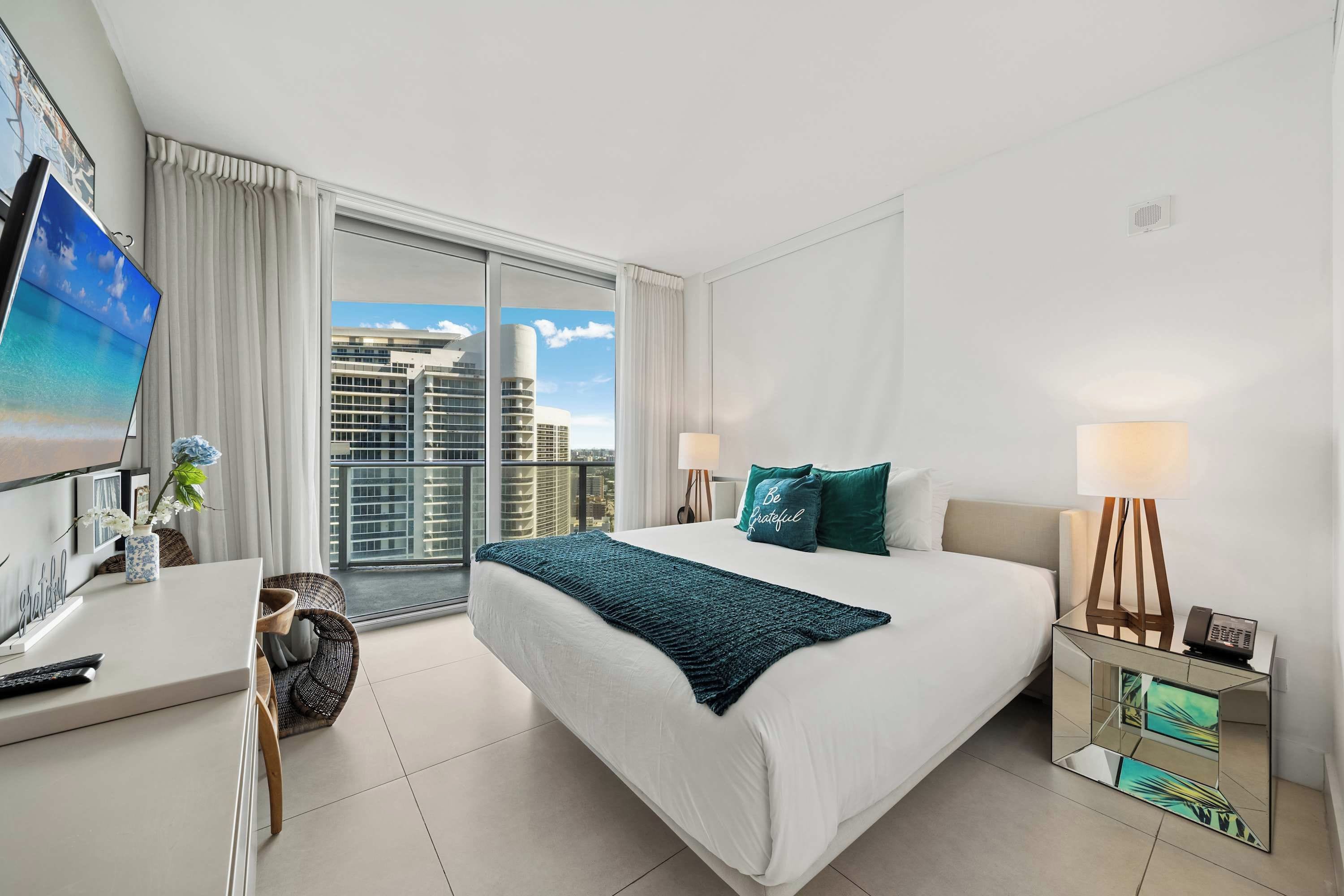 A hotel room with a bed, a desk, a chair, a TV, a window with a city view, and a bedside lamp.