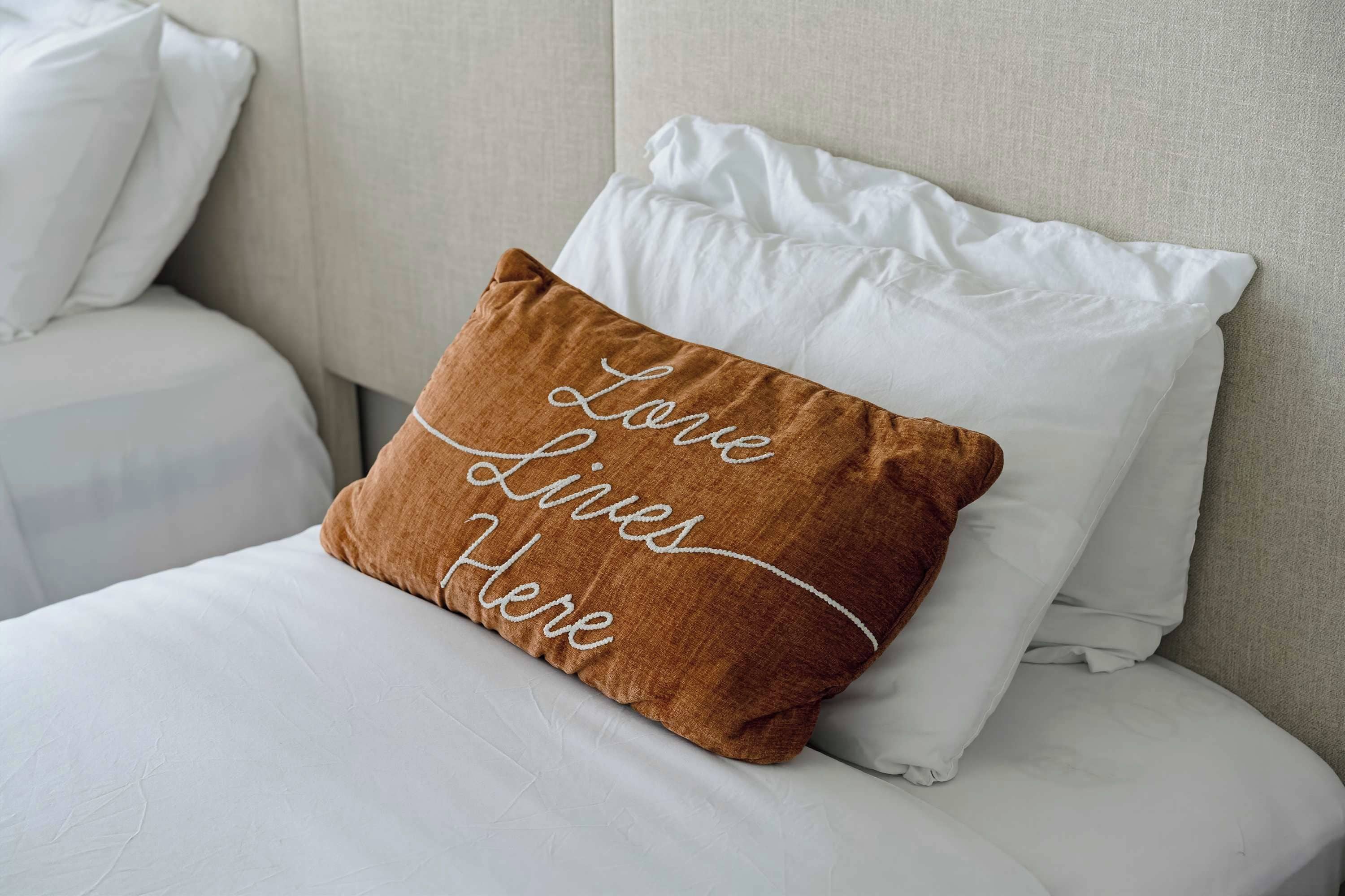 A bed with white bedding and a brown pillow with embroidered text.