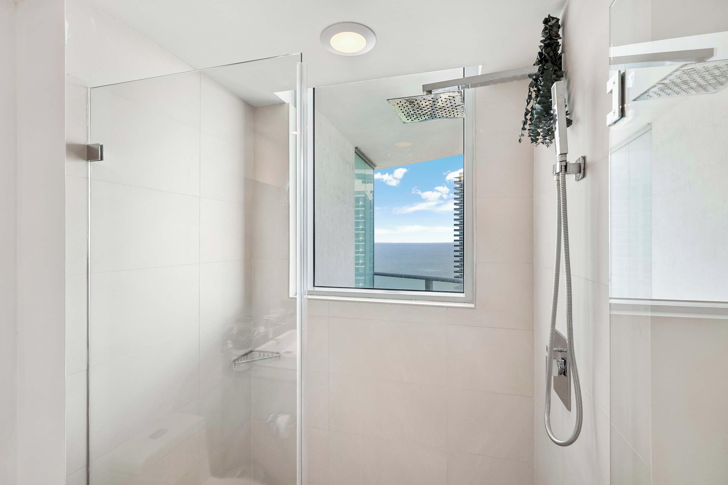 Une douche moderne avec une porte en verre, une fenêtre visible et une vue sur le ciel et les bâtiments.