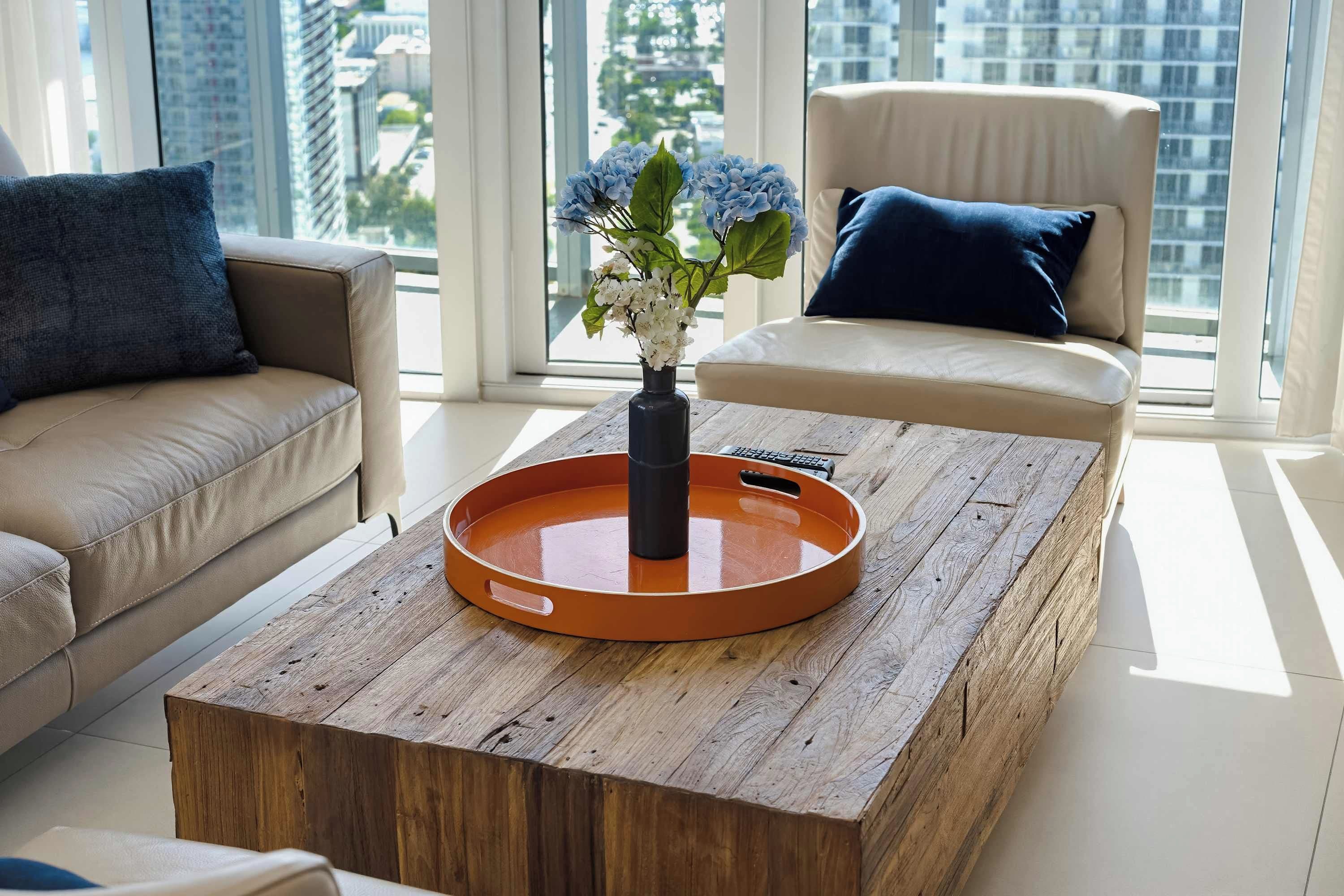 A modern living room with a wooden coffee table, a vase with flowers, a beige sofa, and a chair with a blue pillow.