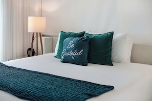 Ein ordentlich bezogenes Bett mit einer türkisfarbenen Tagesdecke, einer Nachttischlampe und zwei grünen Kissen, auf einem davon der Text 'Be Grateful'.