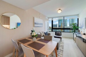 A modern living room with a dining area, large windows with a city view, a round mirror, and a plant. - Sky Corner Residence Pool, Parking, Amenities (Miami)