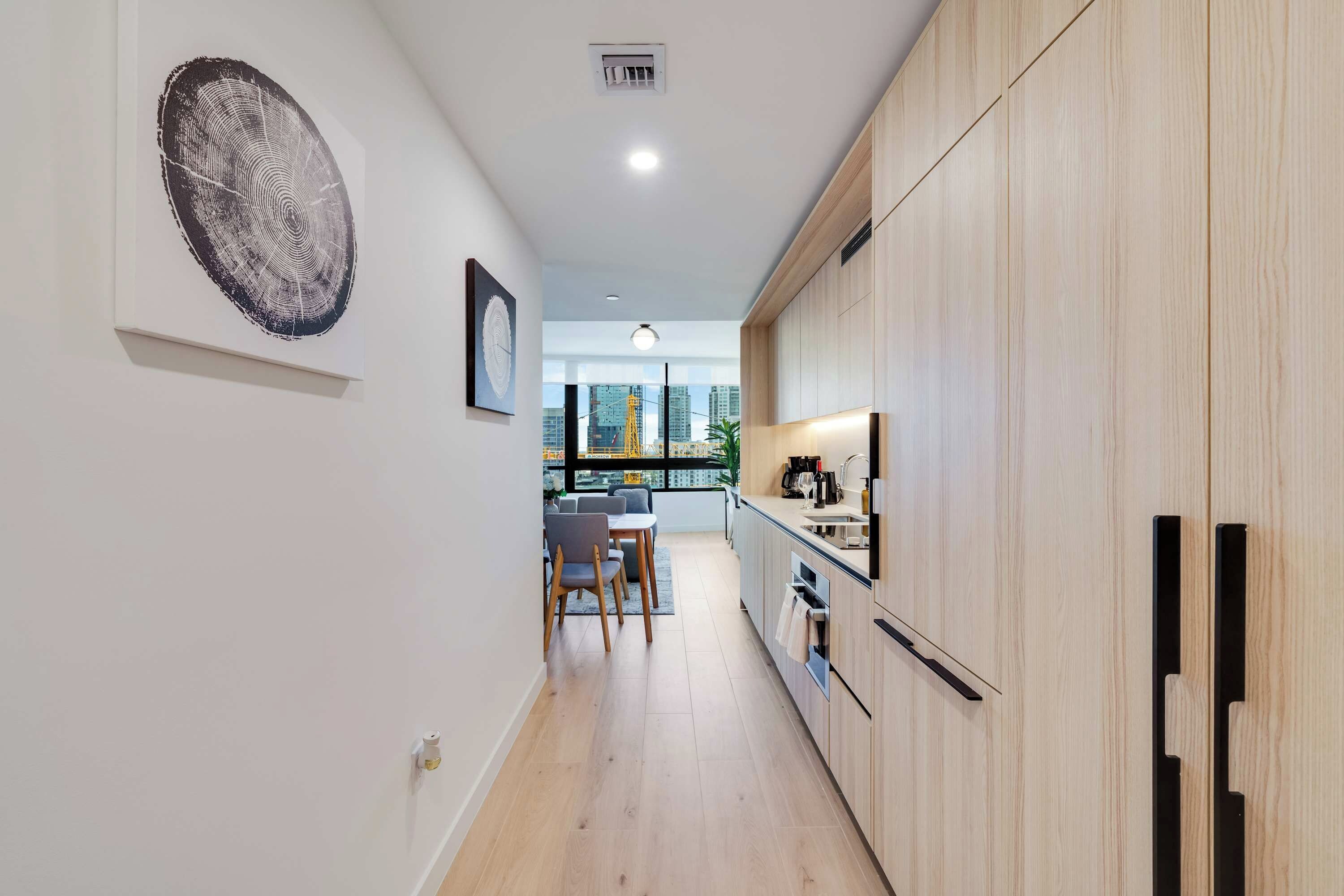 A modern kitchen with wooden cabinets, a dining area with a table and chairs, and a view of the city through large windows.