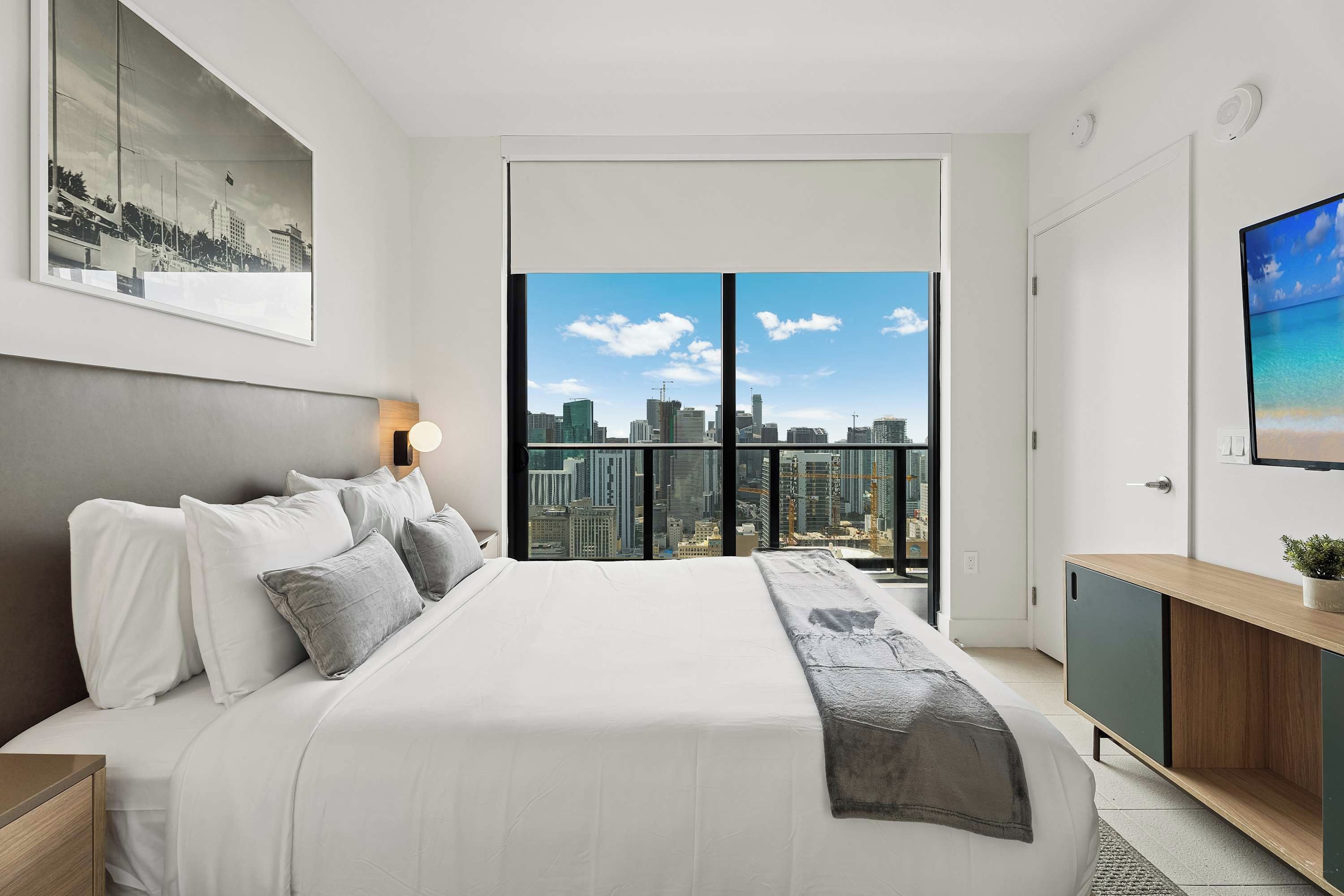 Une chambre moderne avec un grand lit, une vue sur la ville, un tableau encadré au mur et une petite plante sur la commode.