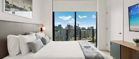 Une chambre moderne avec un grand lit, une vue sur la ville, un tableau encadré au mur et une petite plante sur la commode.
