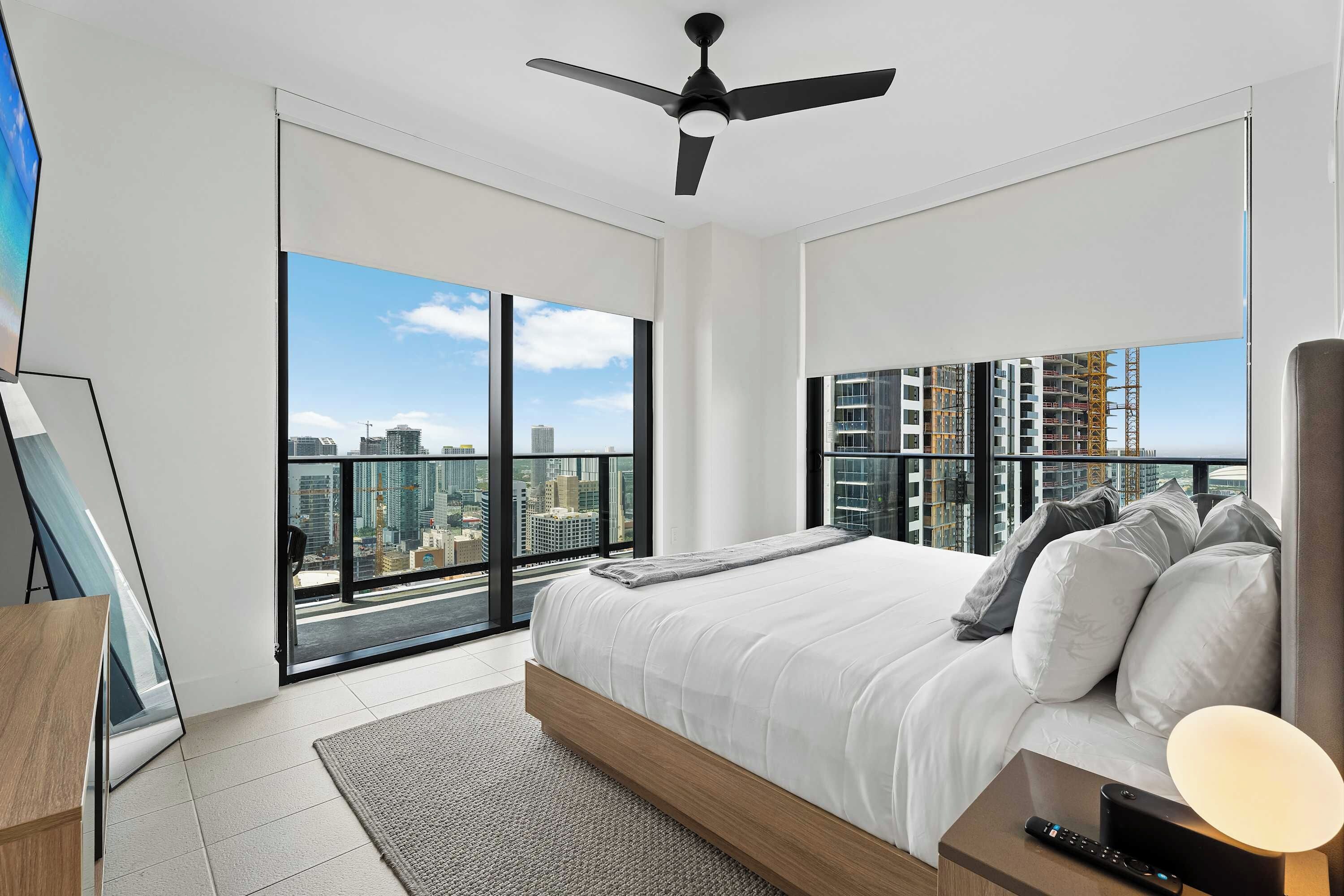 A modern bedroom with a large bed, a ceiling fan, and a city view through floor-to-ceiling windows.