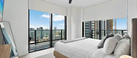 A modern bedroom with a large bed, a ceiling fan, and a city view through floor-to-ceiling windows.