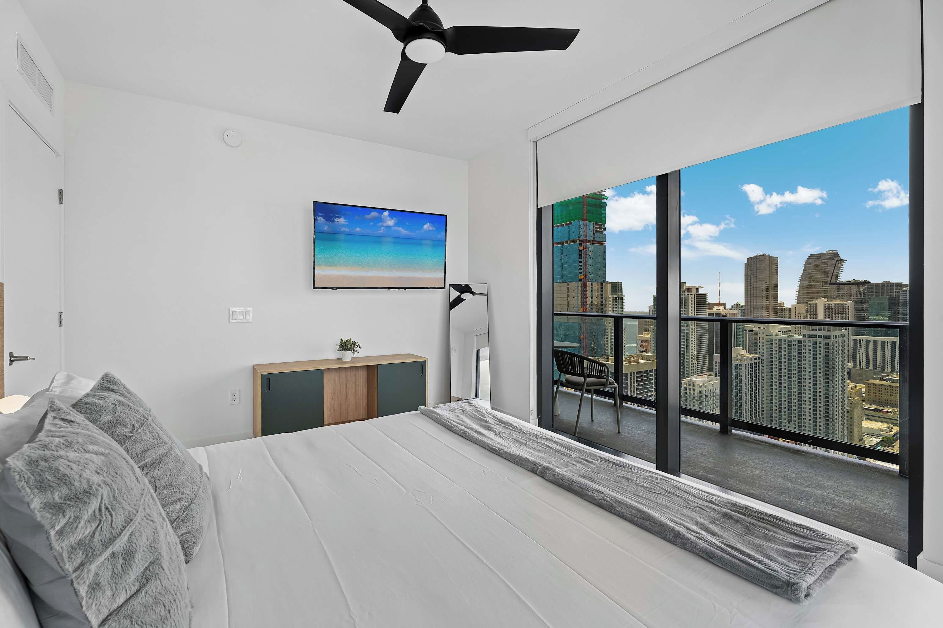 A modern bedroom with a large bed, a ceiling fan, a TV, and a view of a cityscape through large windows.