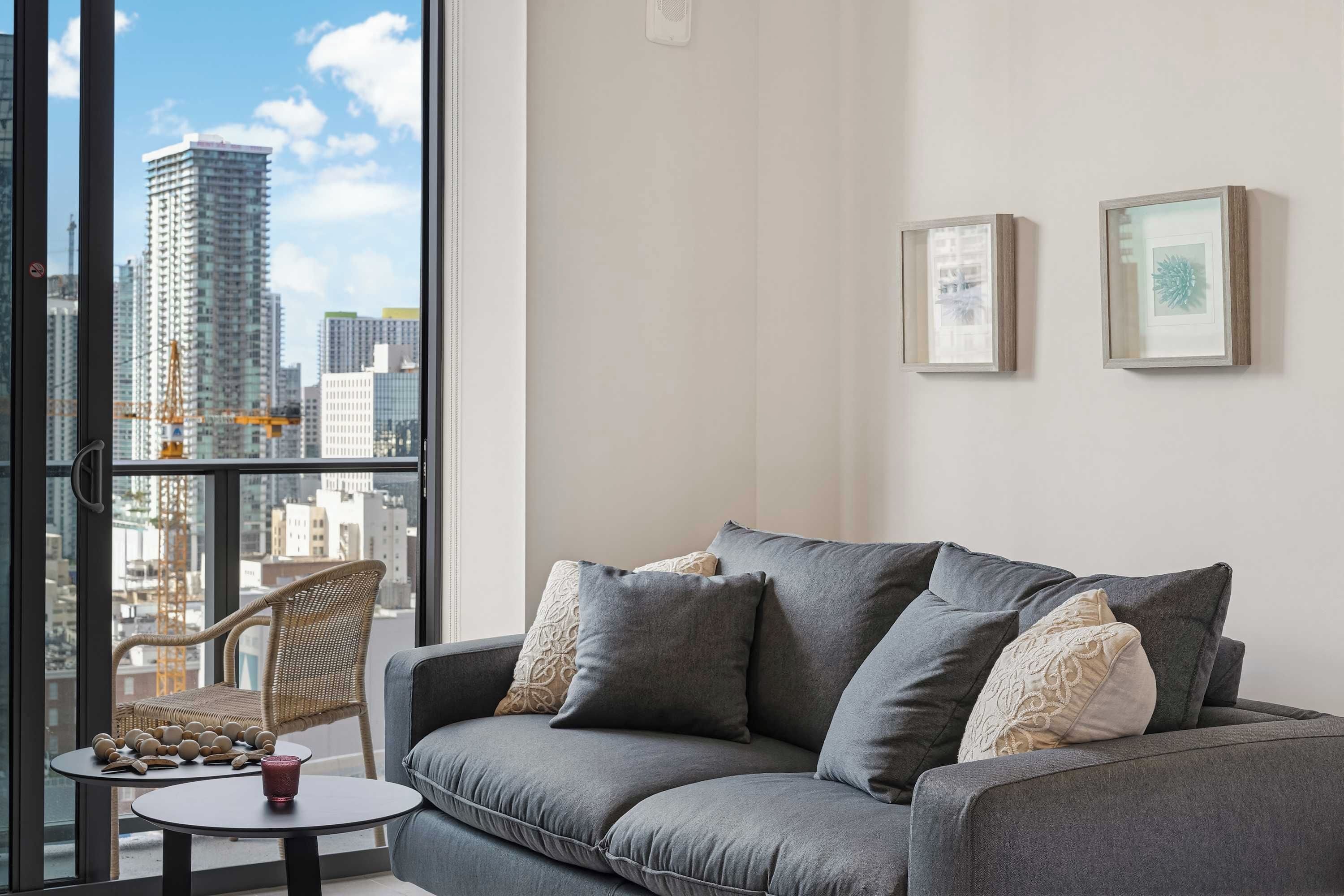 Ein modernes Wohnzimmer mit grauem Sofa, rundem Couchtisch und Korbstuhl. An der Wand hängen zwei gerahmte Bilder und durch große Fenster hat man einen Blick auf die Stadtlandschaft.