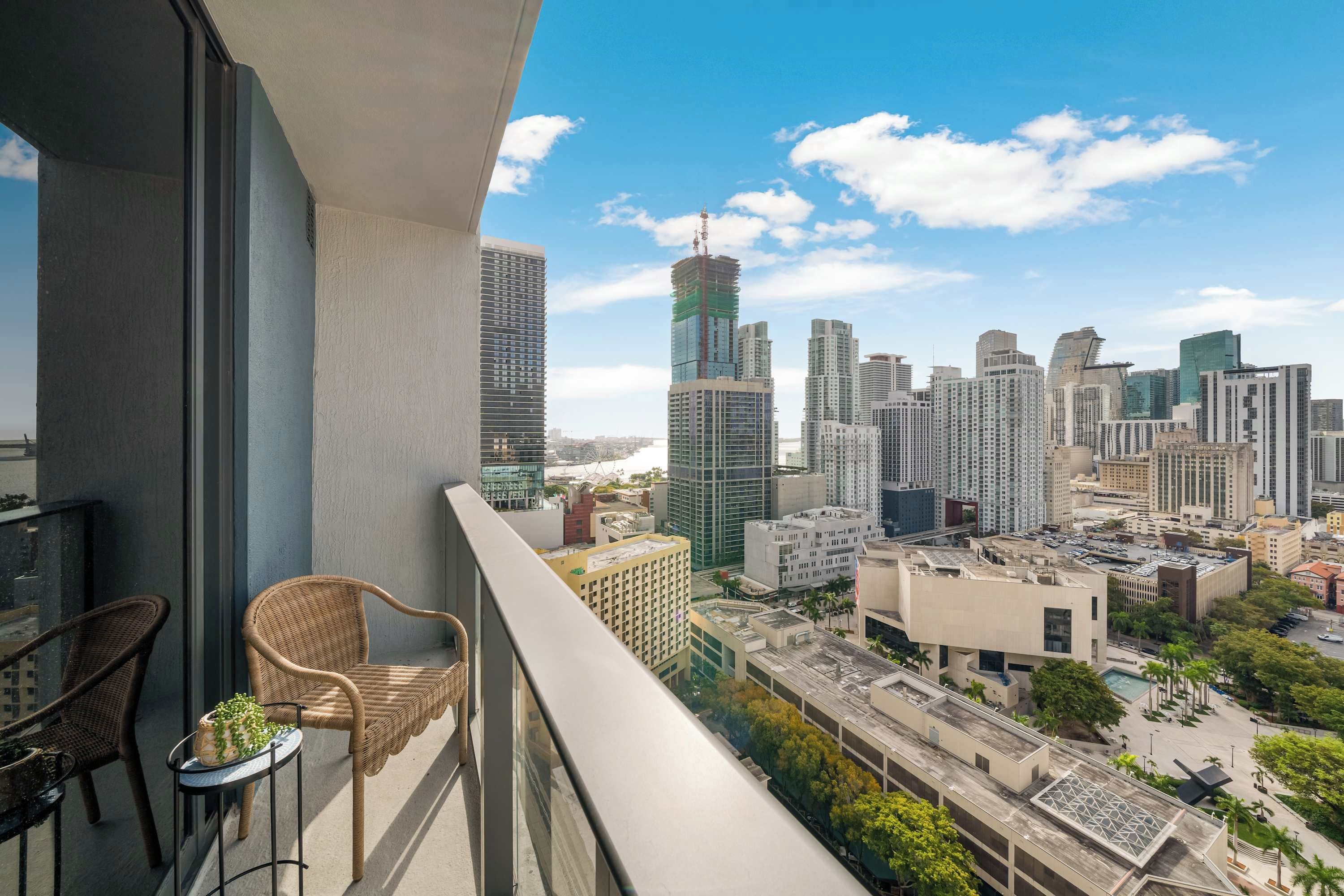 Un balcone con sedie in vimini e un tavolino, con vista su una città caratterizzata da palazzi alti e un cielo sereno.
