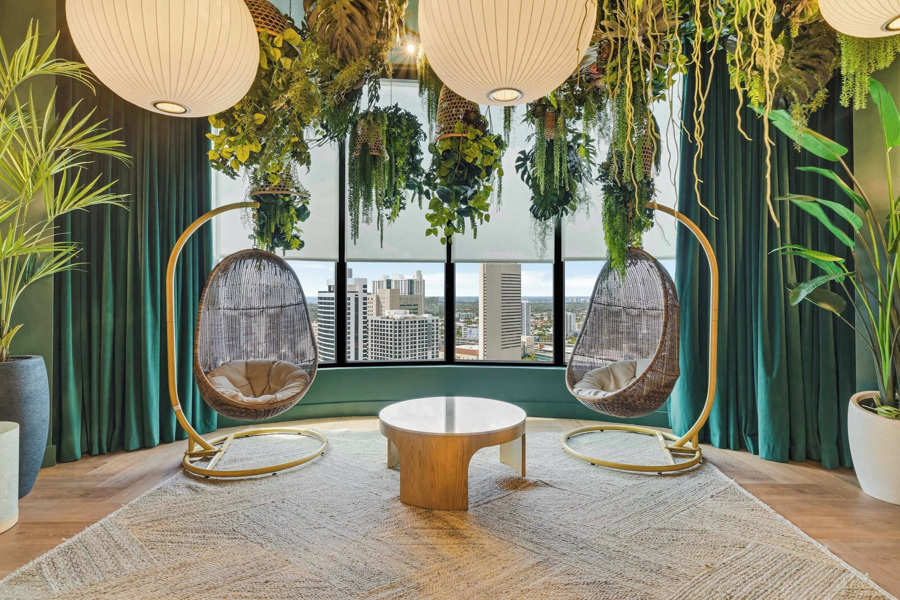 Una habitación con una mesa redonda de madera, dos sillas de mimbre, plantas colgantes y vistas a la ciudad a través de amplios ventanales.