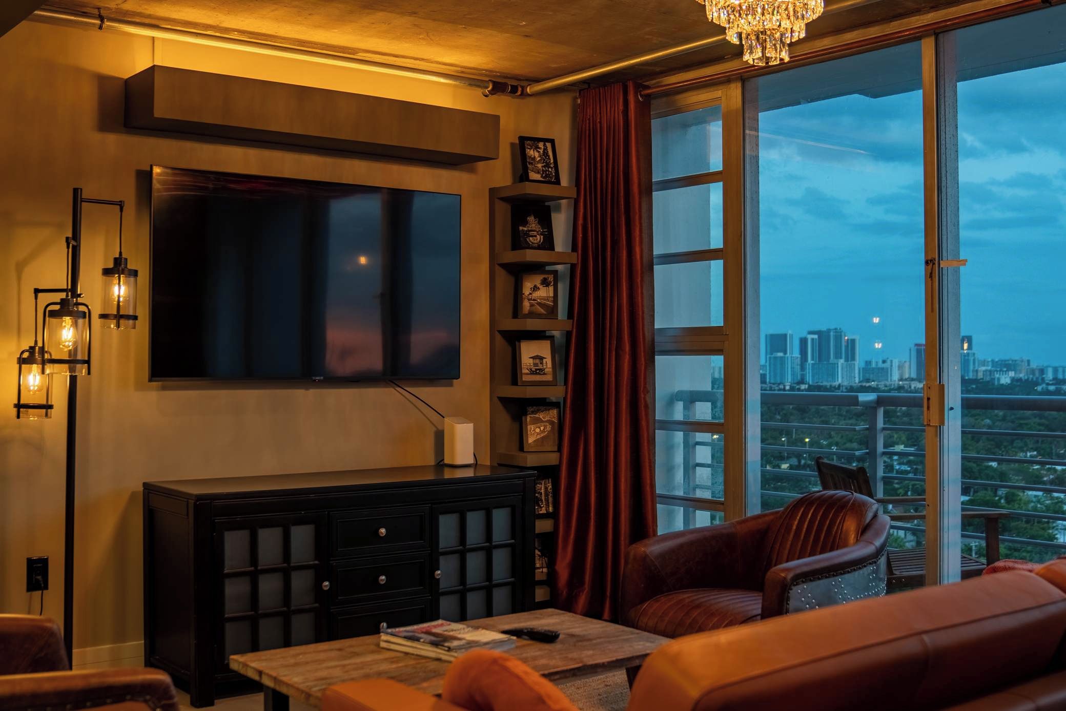 Ein Wohnzimmer mit Flachbildfernseher, Schrank, Ledersesseln, Couchtisch und großem Fenster mit Blick auf die Stadt.
