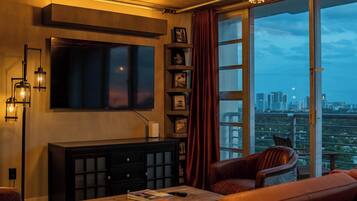 Ein Wohnzimmer mit Flachbildfernseher, Schrank, Ledersesseln, Couchtisch und großem Fenster mit Blick auf die Stadt.